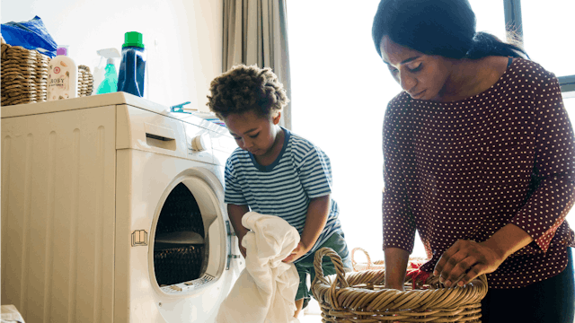 A mom in a brown shirt with white dots and daughter in a blue and white dress taking out laundry fro...