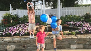 Three boys holding blue and white balloons in the park, while one of them is experiencing a middle-c...