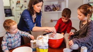 Three kids sitting on and around the table helping their mom, who is the default parent, make food.