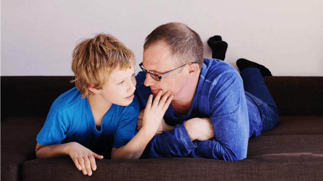 A father in a blue shirt playing with his son in a blue shirt that has autism while lying in a bed