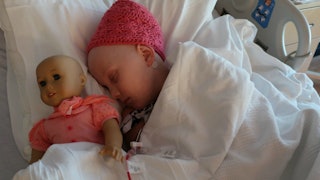 A child with cancer wearing a pink hat, sleeping on the bed while hugging her doll in a pink dress
