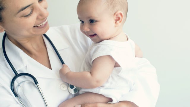 A newborn wearing white clothes smiling while being held in the air by a pediatrician