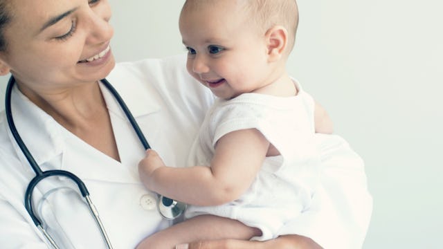 A newborn wearing white clothes smiling while being held in the air by a pediatrician 