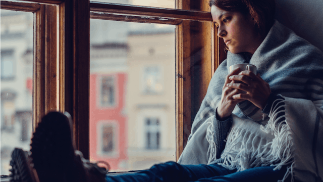 A woman with debilitating anxiety sitting and looking trough the window while wrapped in a blanket a...