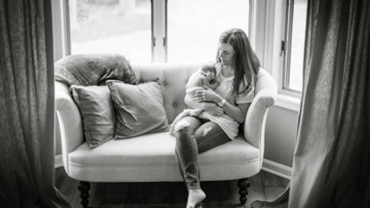 A mother holding her baby in her lap while sitting on a couch next to a window in black and white