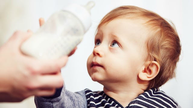 A blonde toddler with blue eyes in a navy shirt with white stripes is reaching for a milk bottle han...
