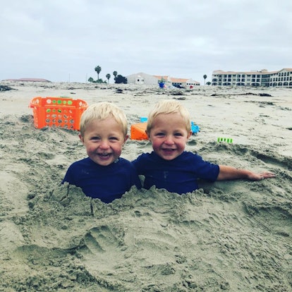 Blonde twin boys wearing blue shirts and smiling while half buried in sand with beach toys behind th...