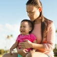 A long-haired brunette woman in a peach dressed shirt holding her crying baby wearing a pink shirt o...