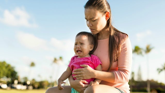 A long-haired brunette woman in a peach dressed shirt holding her crying baby wearing a pink shirt o...