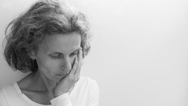 Worried and feared mother in a white shirt with her hand on her cheek looking down and standing alon...