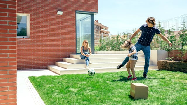 A father playing soccer with his kid in a garden while the mother is sitting and watching them
