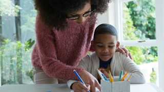 A woman in a pink sweater assists a child in a beige hoodie with his homework