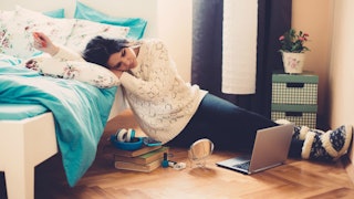 A teen girl sitting on the floor of her room, leaning on a bed, looking at the opened laptop