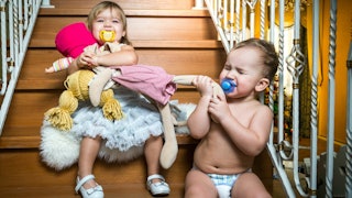 A crying baby and a toddler sitting on stairs, fighting over dolls