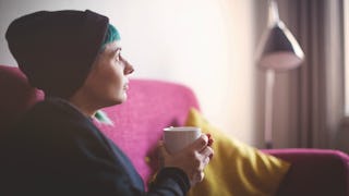 A woman with blue hair in a black sweater and a black beanie holding a white mug and sitting