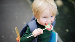 A small boy going to school with a backpack and a blue book and holding a lollipop in his mouth