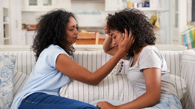 A woman sitting with her teen daughter on the couch, holding her head with one hand, a cup of coffee...