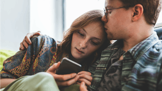 A husband hugging and kissing his wife's forehead while she's looking at her phone