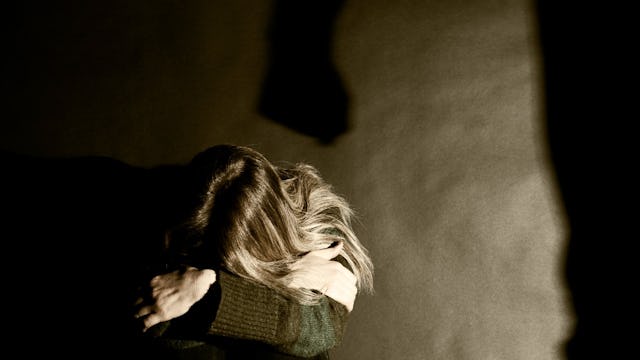 A frightened woman huddled on the floor, her back pressed against the wall, while a dark silhouette ...