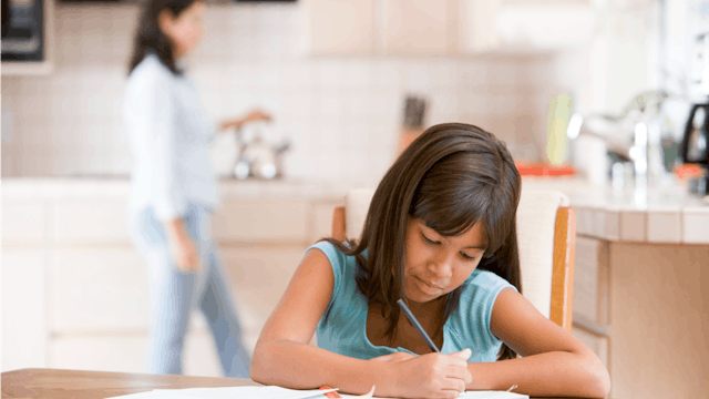 A girl sitting and writing her homework at the table with her mom standing blurred in the background