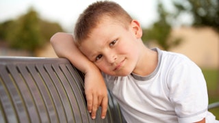 Young boy leaned on a bench, supporting his head with his right arm.