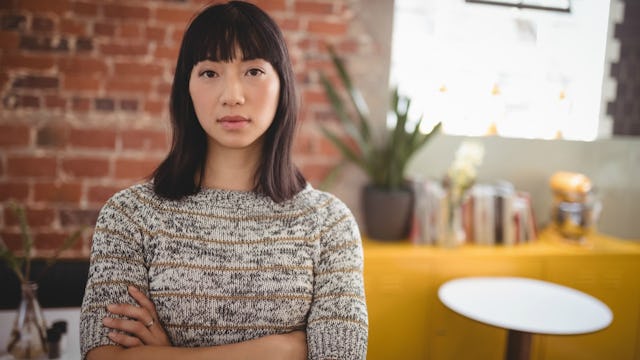A brunette woman in a beige sweater standing in a café with her arms crossed