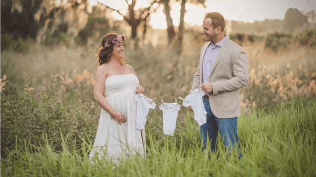 18 weeks triple pregnant woman in a white dress and her husband holding three baby outfits washed an...