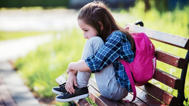 A girl wearing a blue and black plaid shirt, jeans, and a pink backpack curling up on a bench.