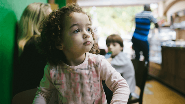 A toddler with curly hair and a pink shirt looking off to the side with two other children behind he...