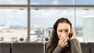 A sick brunette is sitting in the airport terminal, blowing her nose while talking on the phone