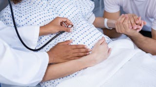 A pregnant woman in a hospital bed with a doctor checking her heart rate and someone holding her han...