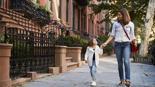 A mom in a white shirt & denim jeans holding her smiling daughter's hand while walking in the street...