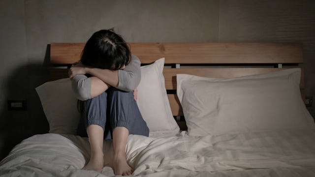 A woman dealing with depression while sitting curled up on a bed in a dark room.