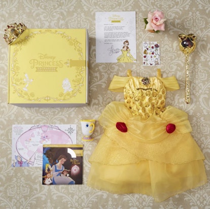 The content of a Disney Princess subscription box next to the box, including a yellow Belle dress, a...