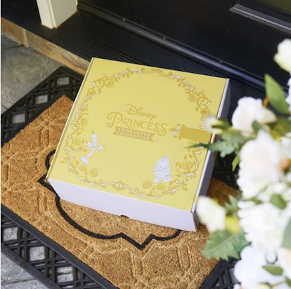 A vanilla yellow Disney Princess subscription box on a doormat next to white roses slightly blurred