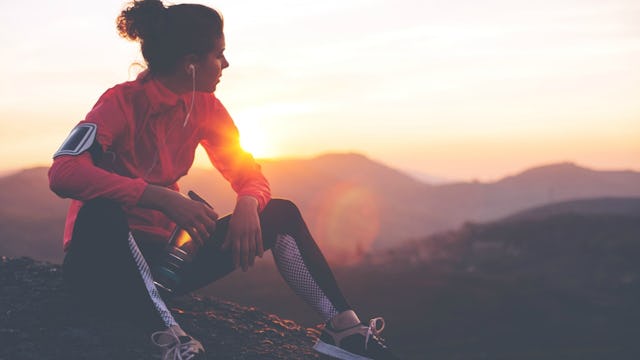A woman wearing an orange sweatshirt and black yoga pants sitting on the top of a hill, watching the...