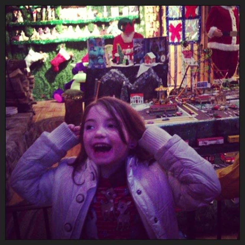 A brown-haired toddler girl standing in front of a Christmas toy stand, smiling brightly, looking ha...