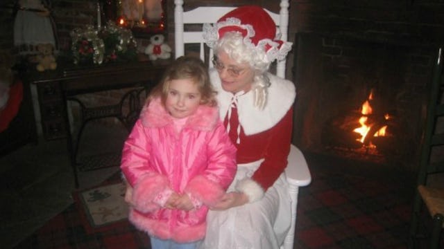 A single mom dressed as Santa, sitting in a white wooden chair, holding closely a blonde toddler gir...