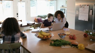 An unhappy child sitting across the mom and other child looking at the laptop