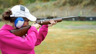 A brown haired teen wearing a white cap and a pink hoodie with shooting range equipment on her aimin...