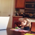 Frustrated mom sitting on the phone in front of her laptop in the kitchen while her daughter is look...