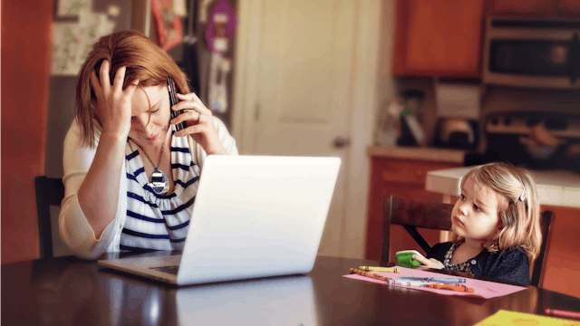 A mom with postpartum rage sitting on the phone in front of her laptop in the kitchen while her daug...