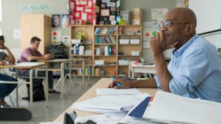 A teacher sitting at his desk in the classroom smiling and looking out at his students as they do a ...