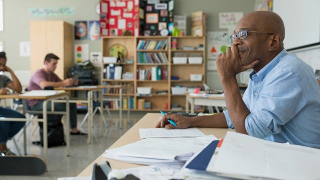 A teacher sitting at his desk in the classroom smiling and looking out at his students as they do a ...