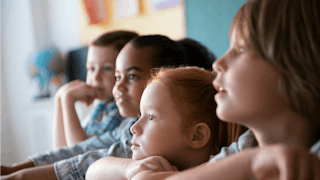 A close-up photo of children in school leaning on their hands against the table and smiling