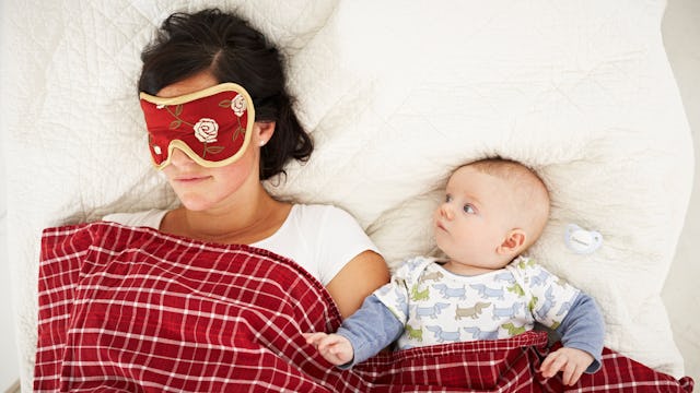 A hungover mom lying down in bed next to her newborn child with a sleep mask covered with a red chec...