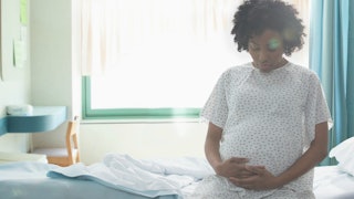 A pregnant woman is sitting on a hospital bed and holding her belly and she has a high risk pregnanc...