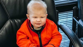 Little toddler boy with DYRK1A sitting down in a leather chair, wearing an orange jacket.