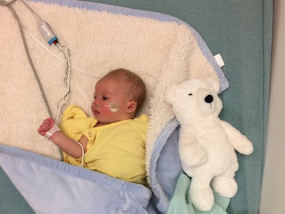 A newborn with RSV, lying down in a yellow shirt wrapped in a blue blanket with a breathing tube wit...