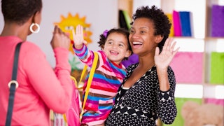 A happy and smiling mother holding a child in her arms as they're waving at a preschool teacher